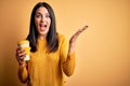 Young woman with blue eyes holding cup of coffee standing over yellow background very happy and excited, winner expression Royalty Free Stock Photo