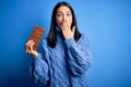 Young woman with blue eyes holding sweet chocolate bar standing over isolated background cover mouth with hand shocked with shame Royalty Free Stock Photo