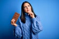 Young woman with blue eyes holding sweet chocolate bar standing over isolated background serious face thinking about question, Royalty Free Stock Photo