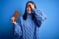 Young woman with blue eyes holding sweet chocolate bar standing over isolated background stressed with hand on head, shocked with Royalty Free Stock Photo