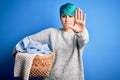 Young woman with blue fashion hair doing domestic chores holding wicker laundry basket with open hand doing stop sign with serious