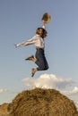 A young woman in blue jeans, shirt and a straw hat jumps on a haystack.