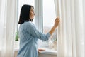Young woman in blue shirt opening curtains looking out the window in the morning in the room, cloudy day in the city Royalty Free Stock Photo