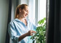 Young woman in blue shirt with spray with water in hands takes care of houseplant in room at home Royalty Free Stock Photo