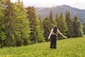 Young woman with a bouquet of flowers and wide open arms standing in the meadow. Forest and mountains in the background. Back view Royalty Free Stock Photo