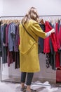 A young woman in a bright coat chooses clothes on a hanger in a store. The pleasure of shopping. Vertical