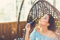 A young woman with bright makeup is sitting in a summer outdoor cafe in a hanging chair and blowing soap bubbles Royalty Free Stock Photo