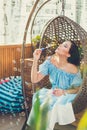 A young woman with bright makeup is sitting in a summer outdoor cafe in a hanging chair and blowing soap bubbles Royalty Free Stock Photo