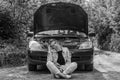 A young woman broke a car on the road and she sits in front of a car with an open hood in confusion and waiting for help with a ca Royalty Free Stock Photo