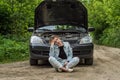 A young woman broke a car on the road and she sits in front of a car with an open hood in confusion and waiting for help with a ca Royalty Free Stock Photo