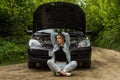 A young woman broke a car on the road and she sits in front of a car with an open hood in confusion and waiting for help with a ca Royalty Free Stock Photo