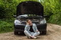 A young woman broke a car on the road and she sits in front of a car with an open hood in confusion and waiting for help with a ca Royalty Free Stock Photo