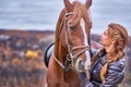 A young woman with brown curly hair caresses a brown horse. The horse caresses the woman.