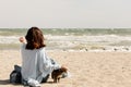Young woman brunette in a light blue cardigan and jeans, with a backpack, sits on the beach and plays with a cat Royalty Free Stock Photo