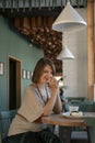 Young woman in cafe with cup of latte or cappuccino and dessert. Flirtatious girl sits at table in cafe