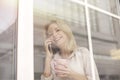 Young woman in cafe standing near window talking on smartphone holding coffee smiling cheerful Royalty Free Stock Photo