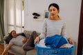 Young woman carrying a basket of dirty clothes doing chores at home while lazy man relaxing and sleeping on sofa couch Royalty Free Stock Photo