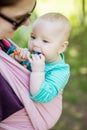 Young woman carrying her baby daughter in woven wrap outdoors in spring park. Baby girl chewing teething beads Royalty Free Stock Photo