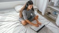 Woman in casual gray dress with headphones using laptop sitting on the bed in the bedroom at home Royalty Free Stock Photo