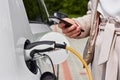 Young woman charging an electric car at public charging station and pays using a mobile phone. Innovative eco-friendly vehicle. Royalty Free Stock Photo