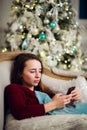 Young woman checking out her text messages on mobile phone as she liyng sofa in front of the Christmas tree at home. Royalty Free Stock Photo