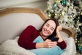 Young woman checking out her text messages on mobile phone as she liyng sofa in front of the Christmas tree at home. Royalty Free Stock Photo