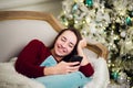 Young woman checking out her text messages on mobile phone as she liyng sofa in front of the Christmas tree at home. Royalty Free Stock Photo