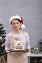 Young woman chef in New Years hat holds gingerbread house in her hands. Vertical frame Royalty Free Stock Photo