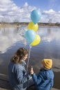 young woman and child are sitting on shore of lake, holding yellow and blue balloons in colors of Ukrainian flag