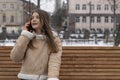 Young woman in city talks on phone and sits on park bench. Girl in beige sheepskin coat rests outside in winter