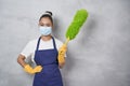 Young woman, cleaning lady in uniform wearing medical mask holding green microfiber duster for cleaning and looking at