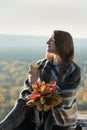 Young woman with closed eyes sits on a hill. Paper cup in hands and a bouquet of yellow leaves. Enjoying a sunny day