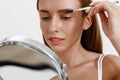 Pretty woman combing her eyebrows with brush and looking at mirror over white studio background Royalty Free Stock Photo