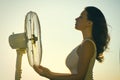 Young Woman cooling herself during hot weather in front of fan on a blue sunset sky background Royalty Free Stock Photo