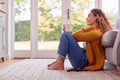 Young Woman In Cosy Warm Jumper Sitting On Floor At Home Looking Out Of Window With Hot Drink