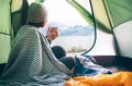 Young woman covered with warm plaid meets cold morning in sitting in a touristic camping tent with a cup of hot tea. Romantic cam