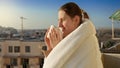 Young woman covering in blanket and enjoying morning sun with cup of tea. People in morning, relaxing at home, enjoying city view Royalty Free Stock Photo