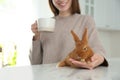 Woman with cup of coffee and adorable rabbit at table indoors, closeup. Lovely pet