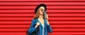 Young woman with cup of coffee wearing black round hat, blue leather jacket and backpack on red background Royalty Free Stock Photo