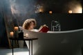 A young woman with curly hair takes a bath in a room with scented candles and reads a book. Beautiful woman is relaxing Royalty Free Stock Photo