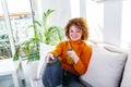 Young woman with curly red hair holding remote control and watching tv, changing channels. Cute girl drinking orange juice and Royalty Free Stock Photo