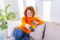 Young woman with curly red hair holding remote control and watching tv, changing channels. Cute girl drinking orange juice and Royalty Free Stock Photo