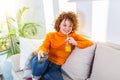 Young woman with curly red hair holding remote control and watching tv, changing channels. Cute girl drinking orange juice and Royalty Free Stock Photo