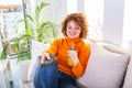 Young woman with curly red hair holding remote control and watching tv, changing channels. Cute girl drinking orange juice and Royalty Free Stock Photo