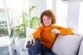 Young woman with curly red hair holding remote control and watching tv, changing channels. Cute girl drinking orange juice and Royalty Free Stock Photo