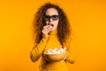 Young woman in 3d glasses watching movie and eating popcorn on yellow background Royalty Free Stock Photo