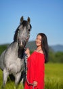 Young woman with dark hair and bright red dress walking her Arabian gray horse in green field on sunny day, smiles happily, Royalty Free Stock Photo