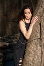 Young woman with dark long hair in black robes is leaning against the tree in the middle of the forest. Back to Nature concept