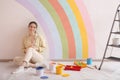 Young woman and decorator`s tools near wall with painted rainbow