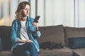 Young woman in denim shirt sitting at home on couch and using smartphone,nearby lies cat. Girl uses digital gadget. Royalty Free Stock Photo